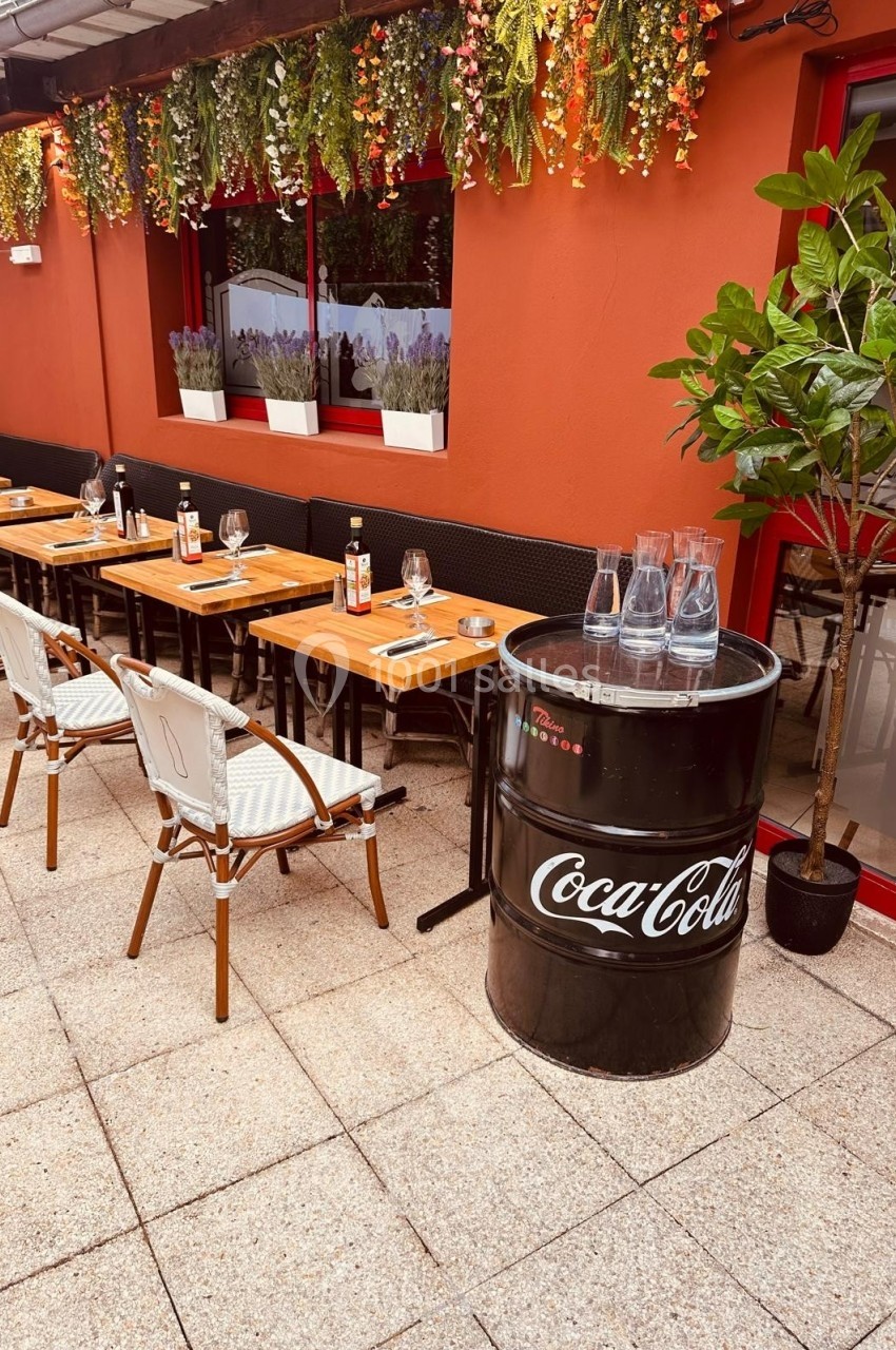 Terrasse d'un restaurant avec tables en bois, chaises blanches, plantes suspendues et un tonneau noir marqué Coca-Cola.