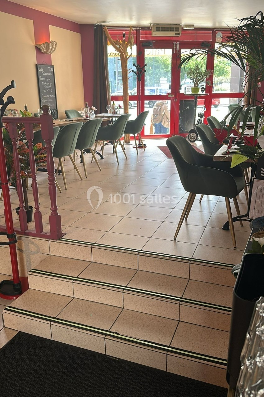 Salle de restaurant lumineuse avec tables dressées, chaises vertes et escalier menant à une entrée vitrée.