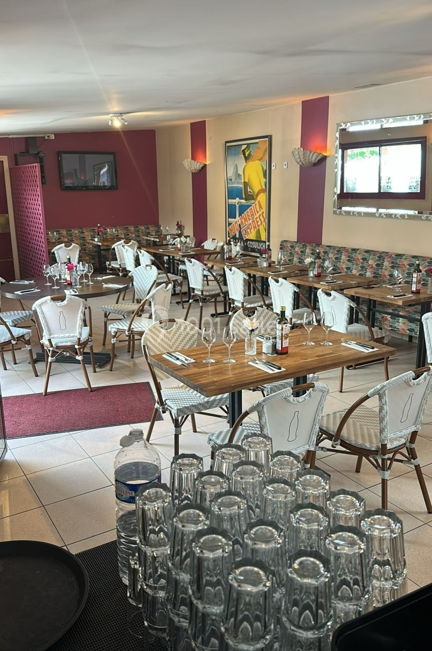 Salle de restaurant avec tables dressées, chaises en rotin, affiches murales et lumière naturelle entrant par une fenêtre.