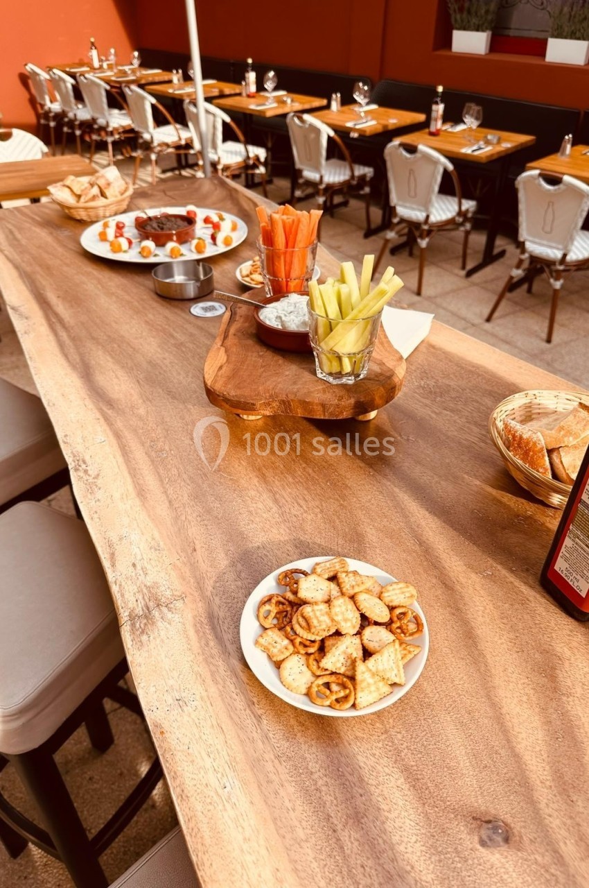 Table en bois avec apéritifs variés, dont légumes croquants, roulés salés et brochettes, en terrasse d'un restaurant.