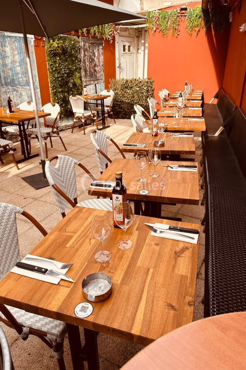 Terrasse ensoleillée d'un restaurant avec tables en bois dressées, chaises blanches et murs rouges ornés de plantes.