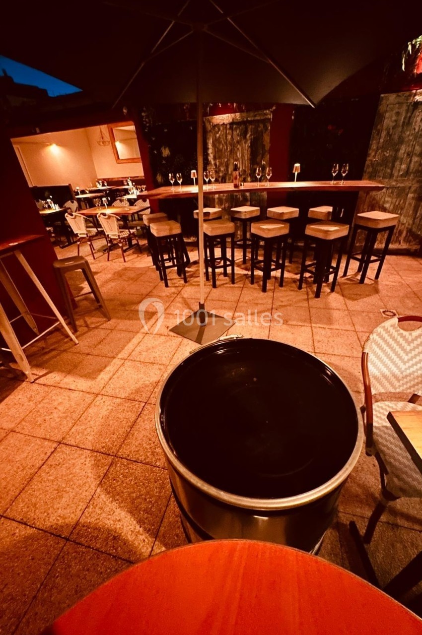 Terrasse extérieure d'un bar en soirée, avec tables, chaises hautes et parasol sous un éclairage tamisé.