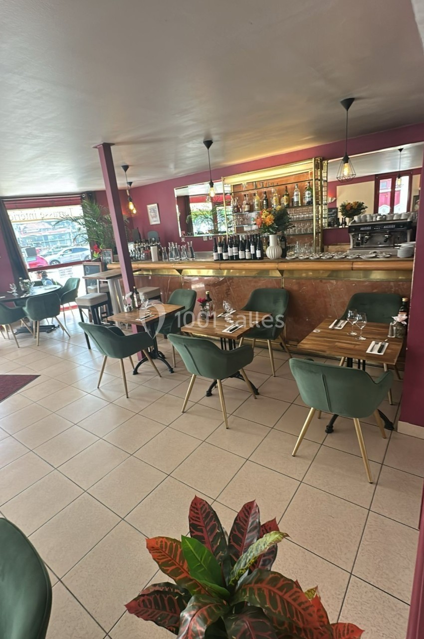 Salle de restaurant lumineuse avec tables en bois, chaises vertes, bar en arrière-plan et plantes décoratives.