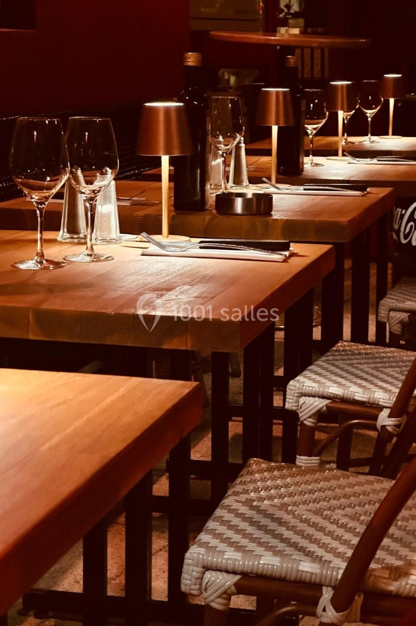 Tables en bois alignées avec chaises en osier, verres à vin et petites lampes dans une ambiance tamisée.