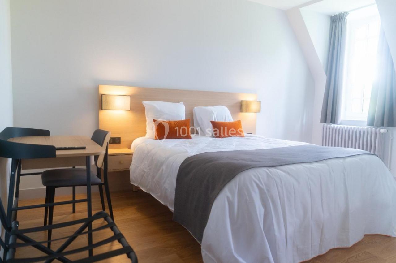 Chambre lumineuse avec un lit double, des coussins orange, un bureau en bois clair et une fenêtre avec rideaux gris.