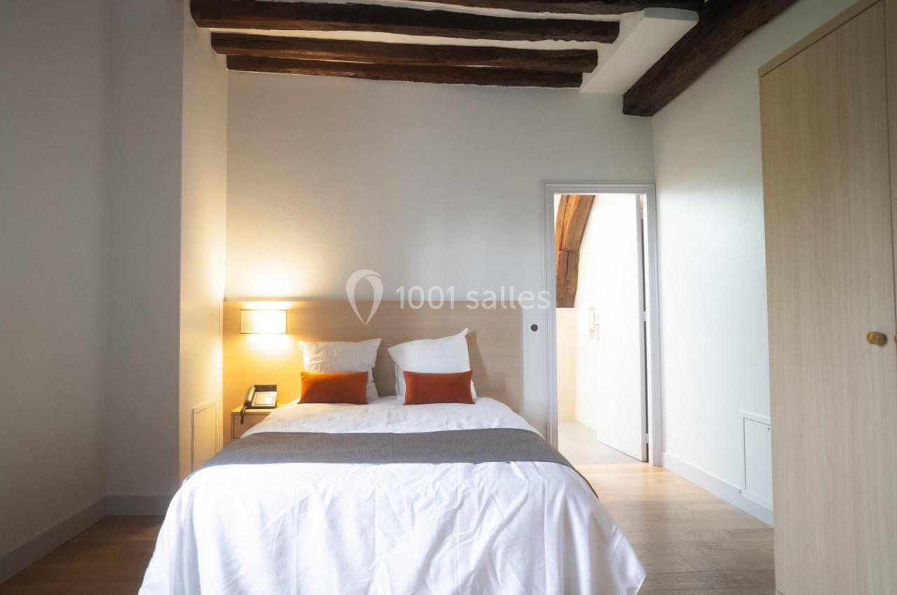 Chambre lumineuse avec lit double, poutres apparentes au plafond, armoire en bois et porte ouverte sur une autre pièce.