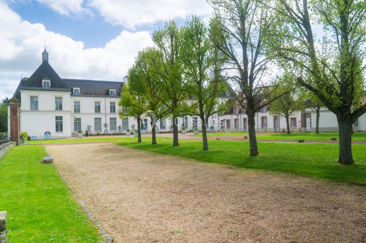 Allée gravillonnée bordée d'arbres menant à un bâtiment historique blanc sous un ciel partiellement nuageux.
