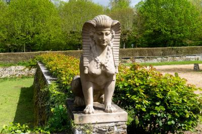 Statue de sphinx en pierre posée sur un socle, entourée de végétation et d'une haie dans un jardin.