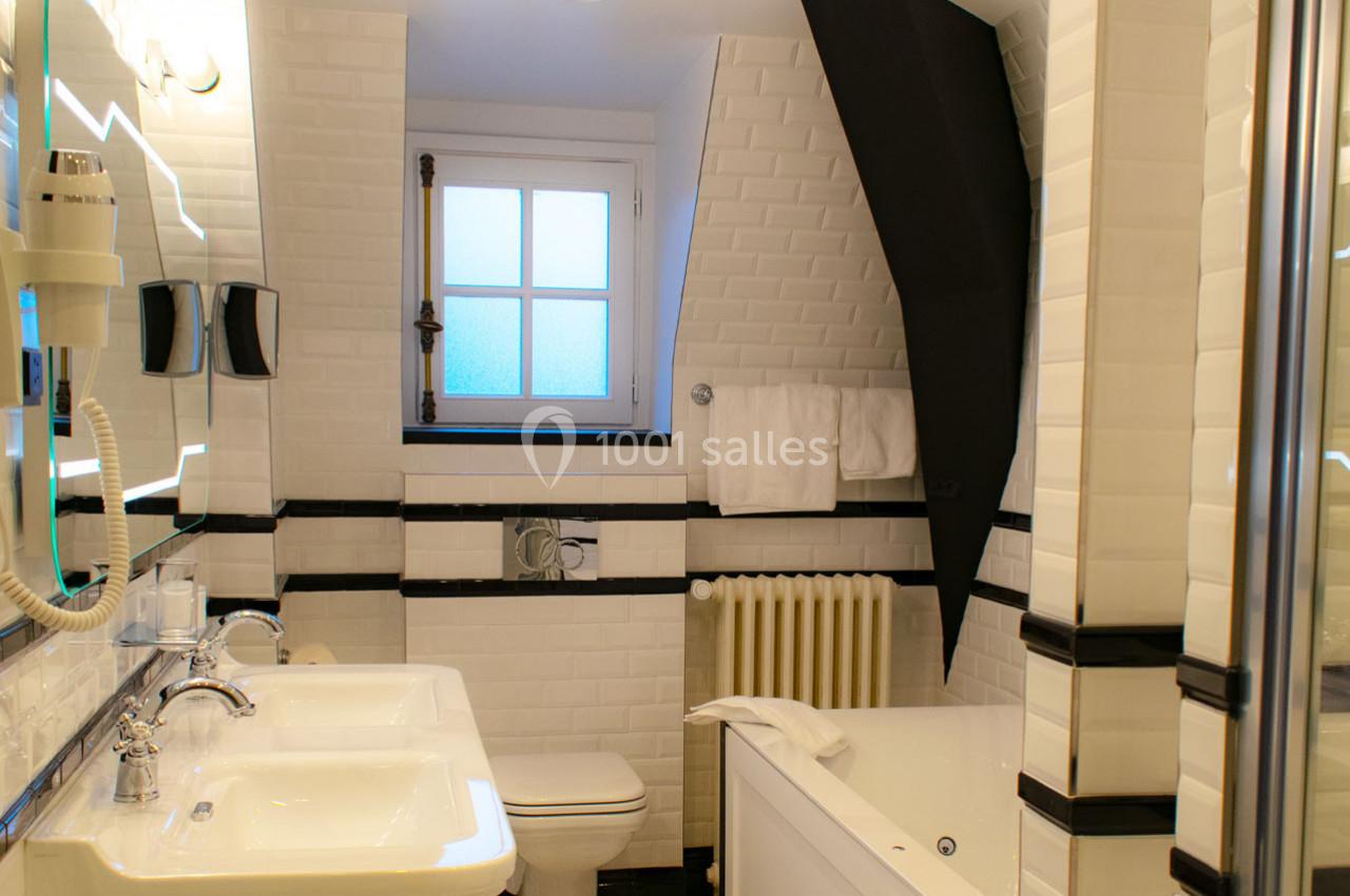 Salle de bain lumineuse avec double vasque, baignoire, toilettes et murs en carrelage blanc avec détails noirs.