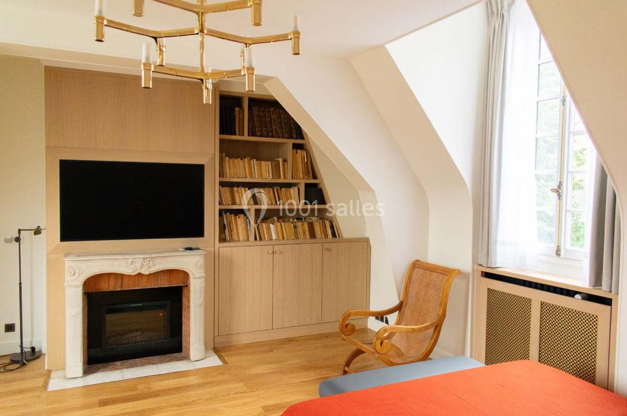Chambre lumineuse avec parquet, cheminée décorative, étagères remplies de livres et fauteuil en osier près d'une fenêtre.