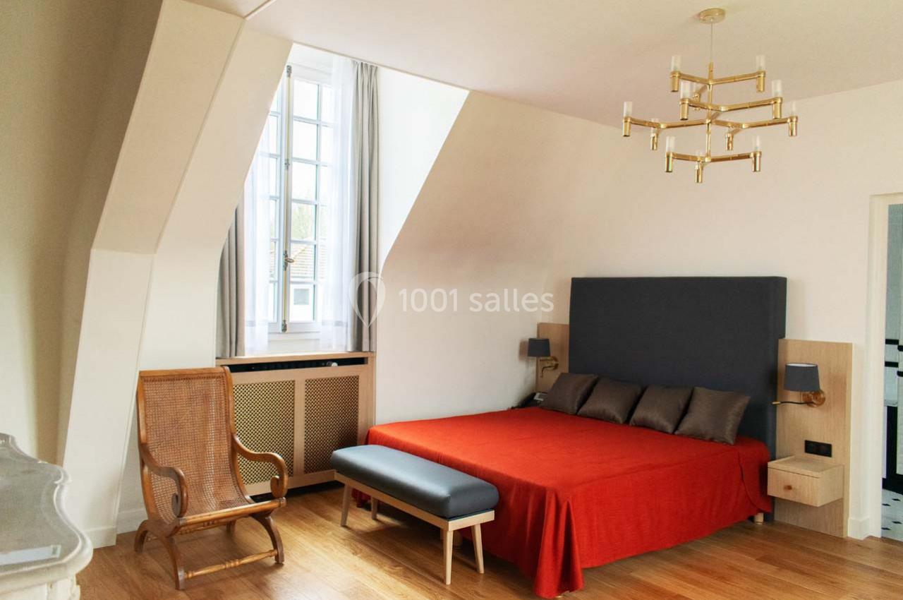 Chambre lumineuse avec lit double à drap rouge, fauteuil en bois, banc au pied du lit et fenêtre mansardée.