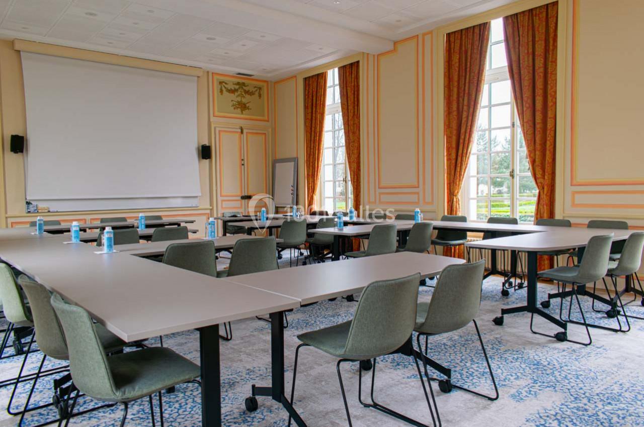 Salle de réunion lumineuse avec tables en U, chaises modernes, écran de projection et grandes fenêtres donnant sur un jardin.