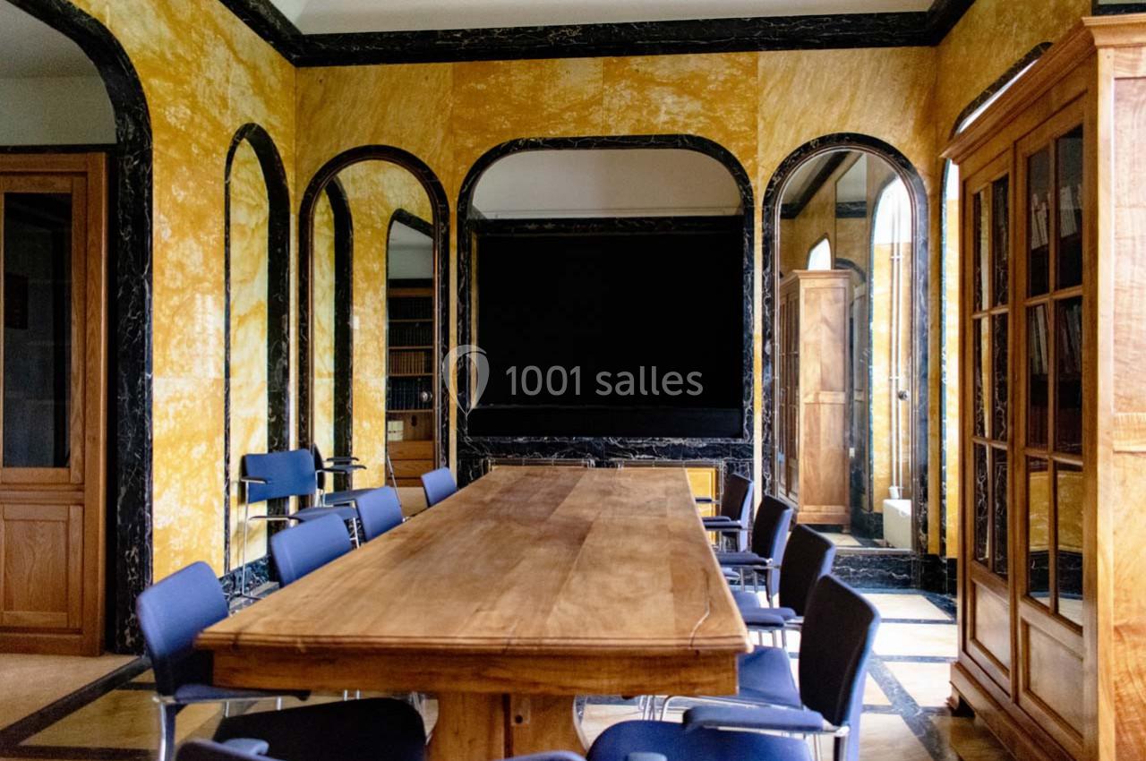 Salle de réunion avec table en bois, chaises bleues, murs jaunes texturés et grandes portes vitrées.