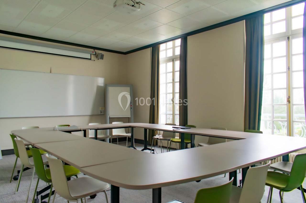 Salle de réunion lumineuse avec tables disposées en U, chaises vertes et blanches, tableau blanc et grandes fenêtres.