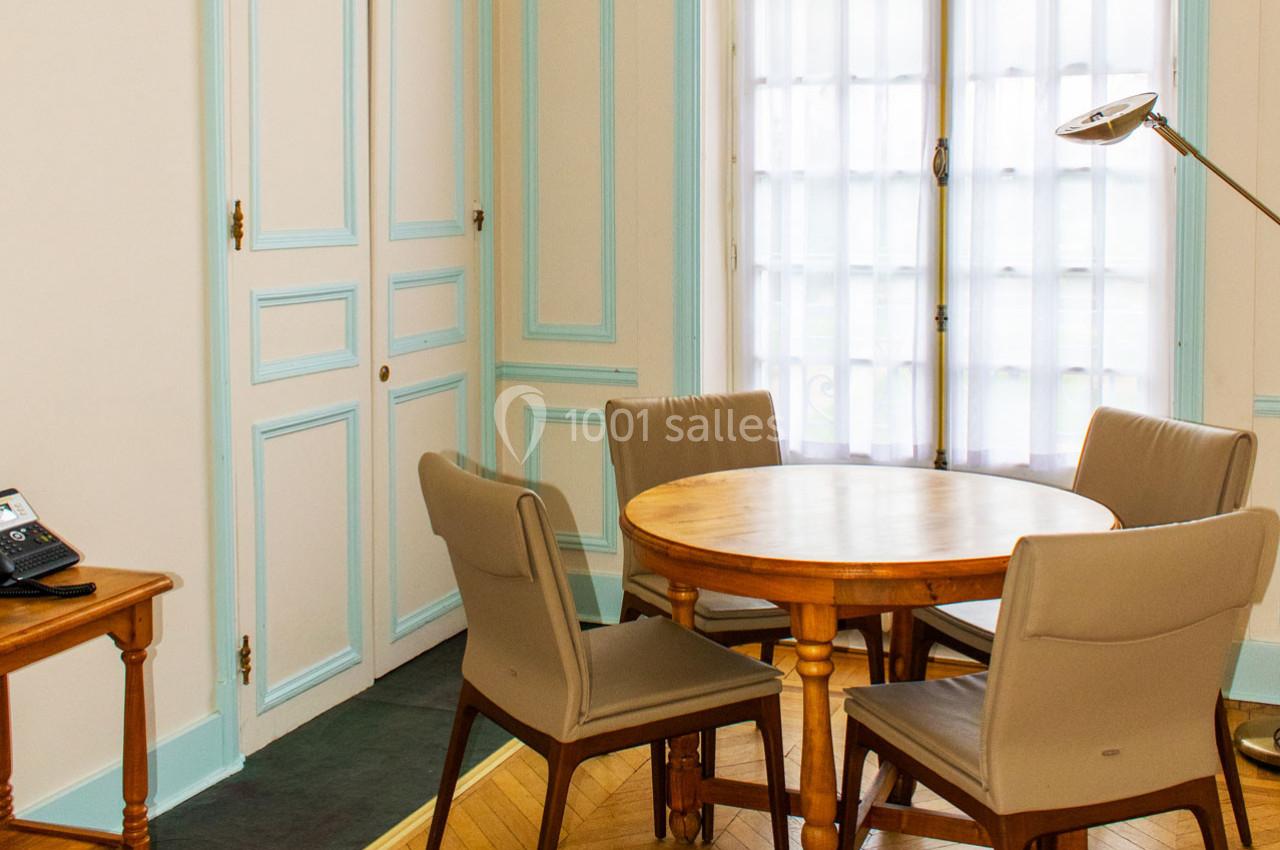 Salle lumineuse avec une table ronde en bois entourée de quatre chaises, près d'une fenêtre avec rideaux translucides.