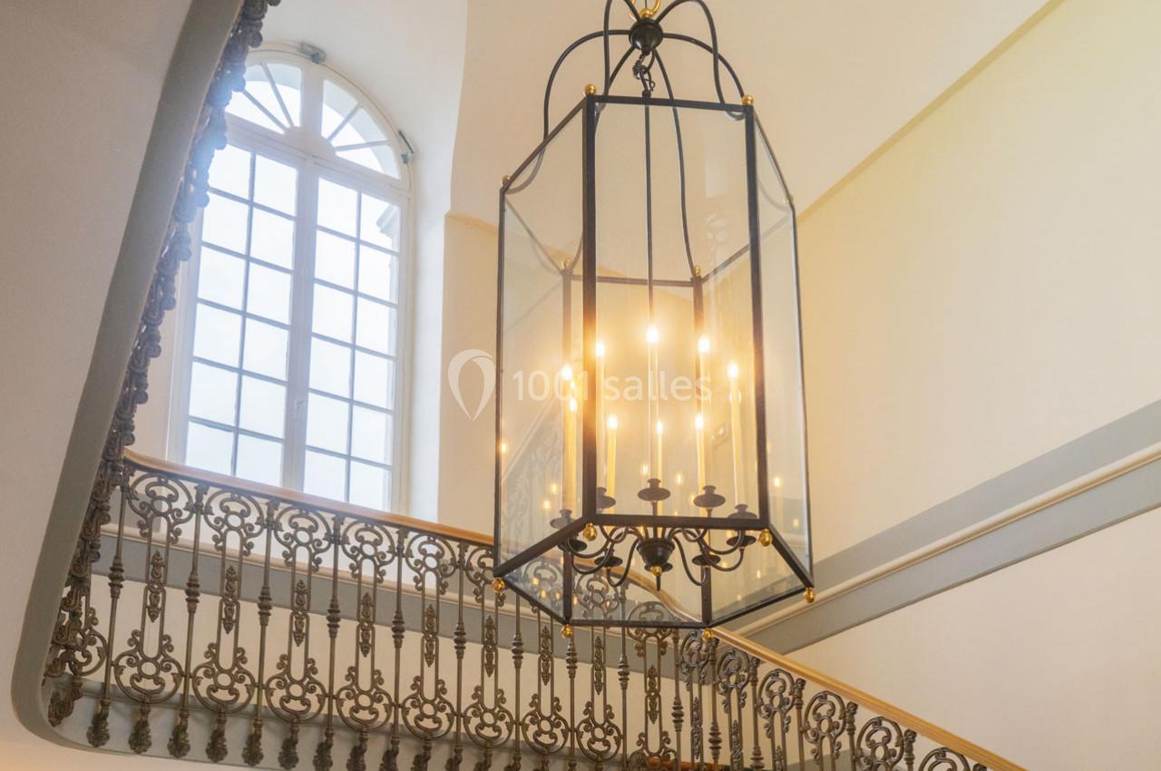 Lustre suspendu en métal noir avec bougies électriques, éclairant un escalier orné de balustrades en fer forgé.