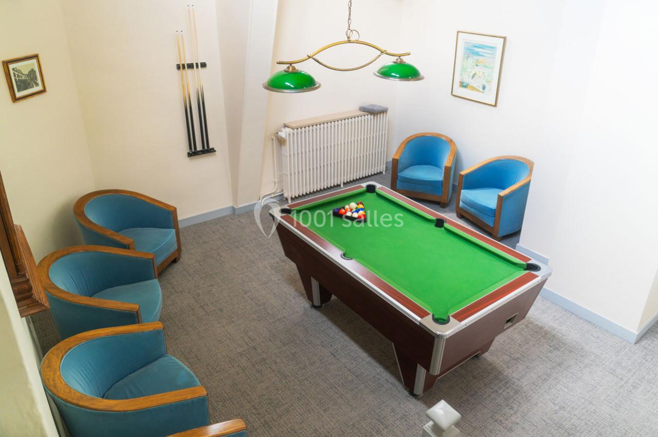 Salle avec une table de billard, quatre fauteuils bleus, un porte-queues mural et des luminaires suspendus verts.