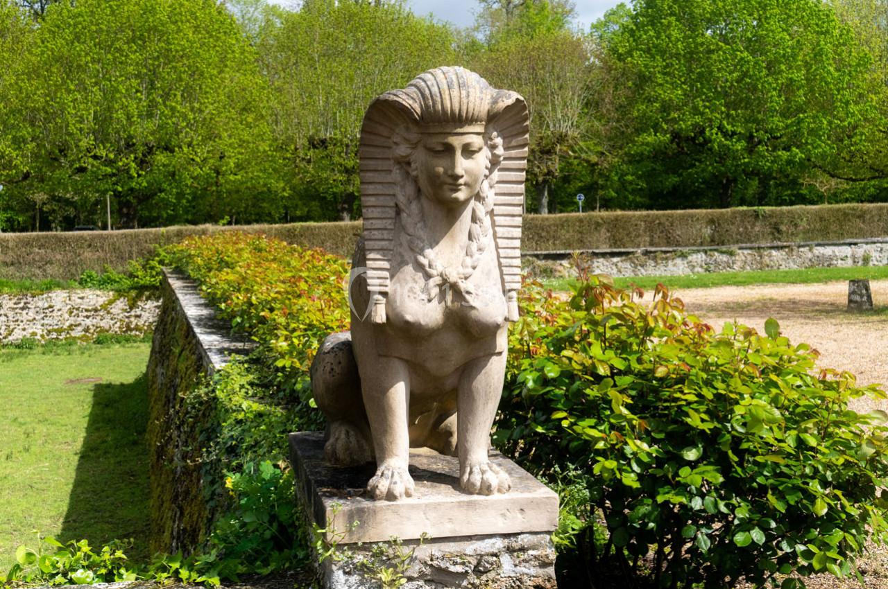 Statue de sphinx en pierre posée sur un socle, entourée de verdure dans un jardin.