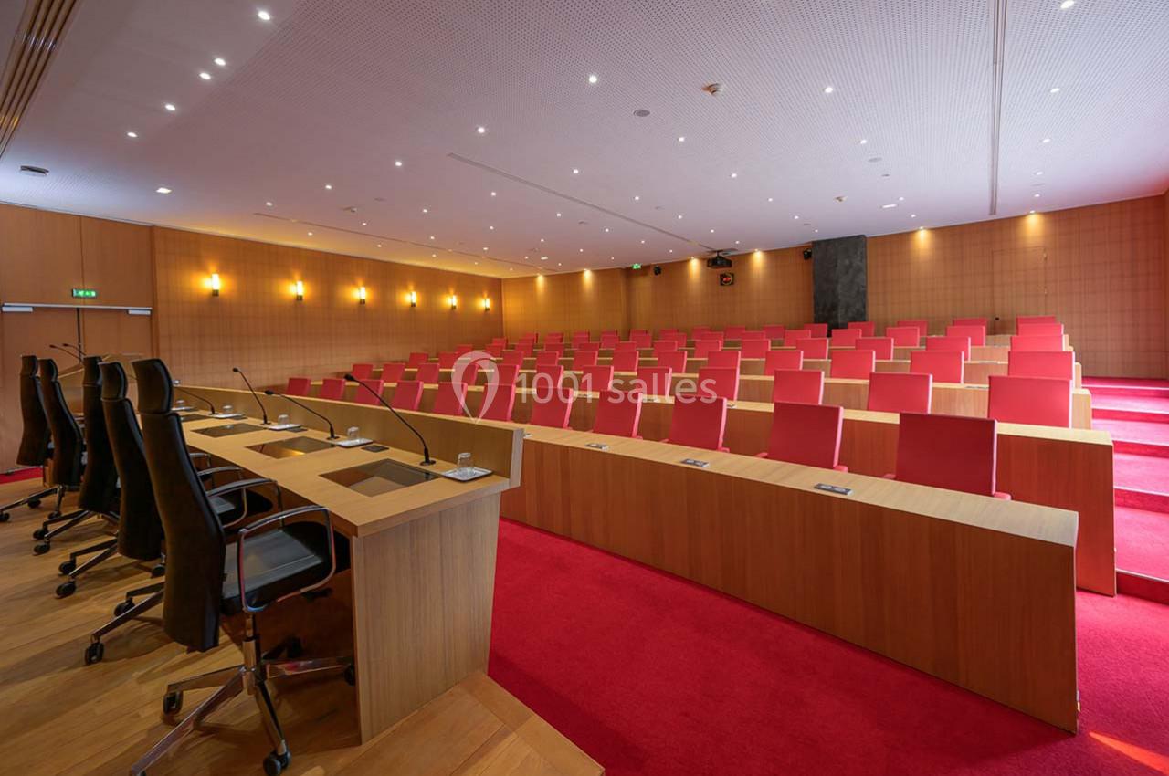 Salle de conférence avec rangées de sièges rouges, pupitres en bois équipés de micros et éclairage au plafond.