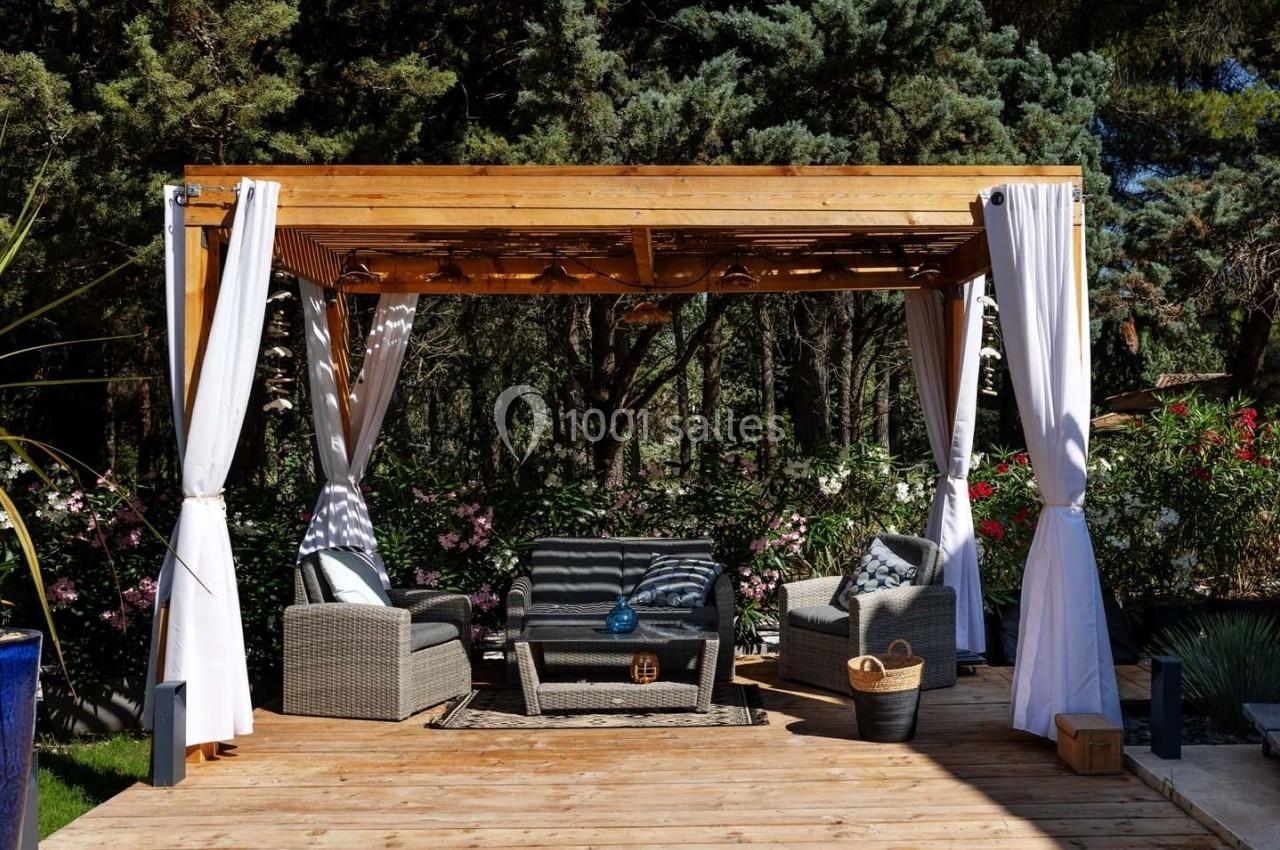 Pergola en bois avec rideaux blancs, mobilier de jardin en rotin et végétation luxuriante en arrière-plan.