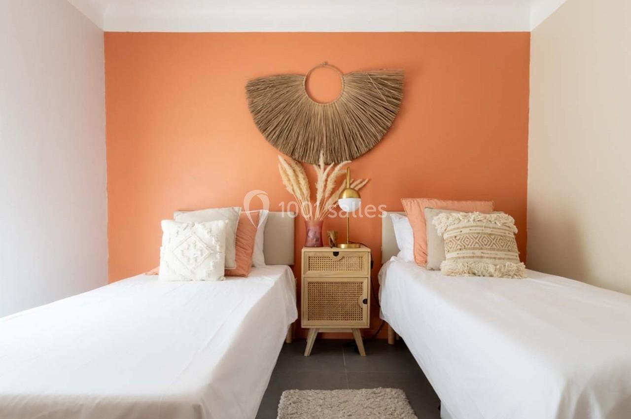 Deux lits simples avec des coussins décoratifs, une table de chevet en rotin et un mur orange orné d'une décoration murale.