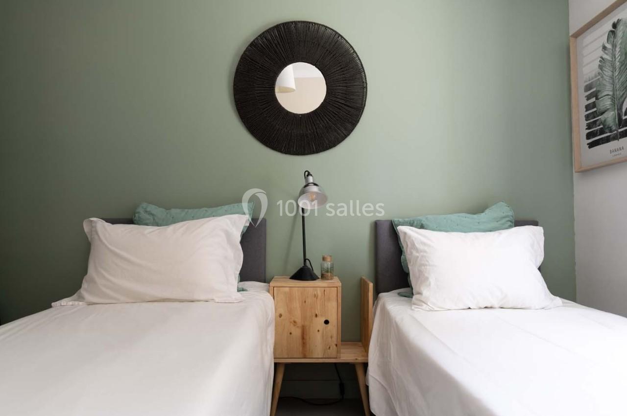 Deux lits simples avec des draps blancs, une table de chevet en bois, une lampe et un miroir rond sur un mur vert.