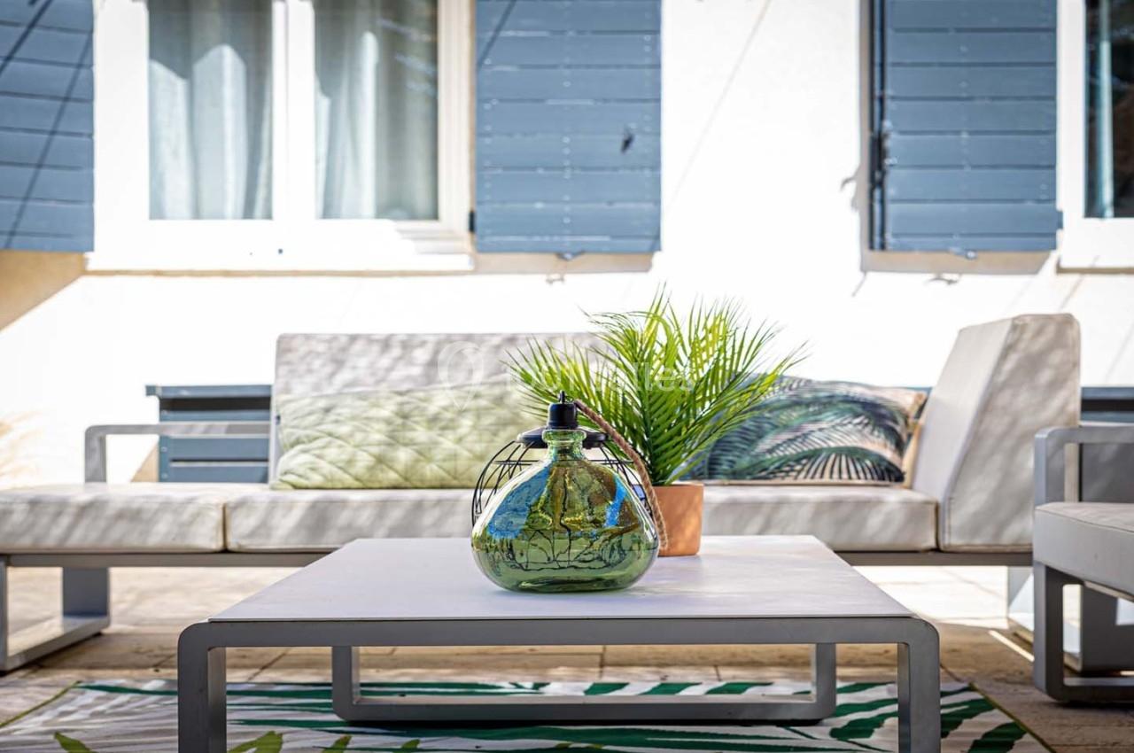 Table basse avec une bonbonne en verre et une plante, entourée de fauteuils sur une terrasse ensoleillée.