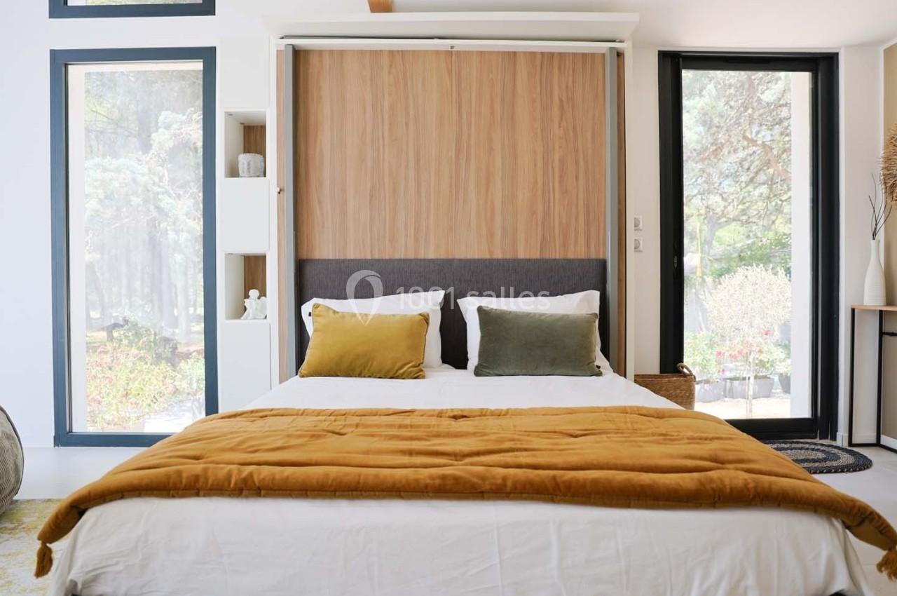 Lit double avec une couverture ocre dans une chambre lumineuse, dotée de grandes fenêtres et d'un décor minimaliste.