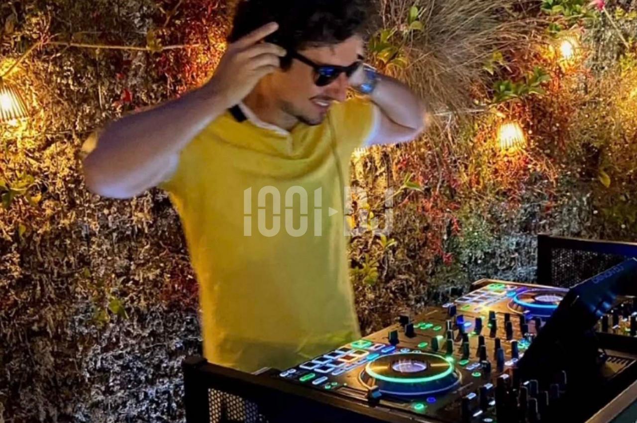 Un DJ en chemise jaune mixe de la musique sur une console éclairée, entouré de lumières et de végétation.
