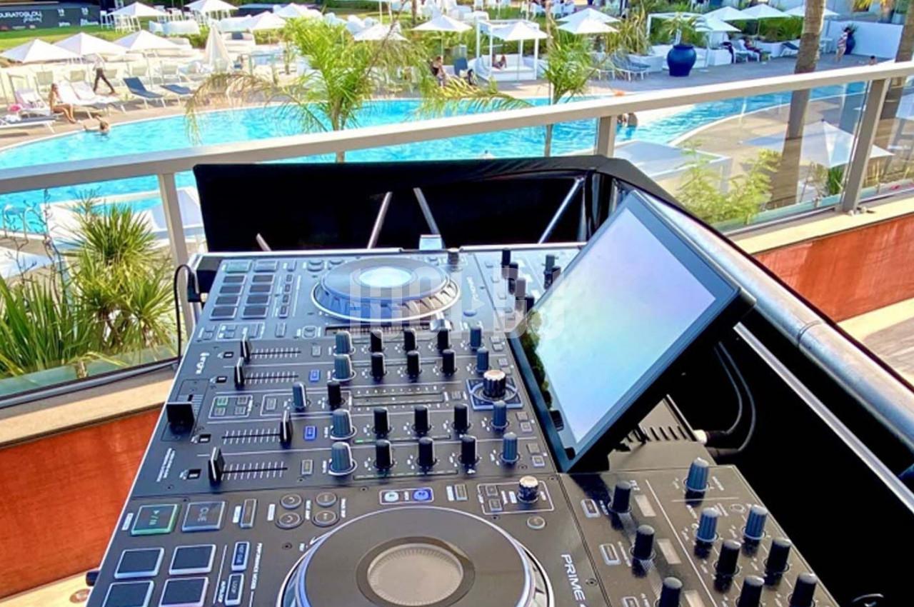 Table de mixage DJ installée en extérieur, avec vue sur une piscine entourée de transats et de parasols.