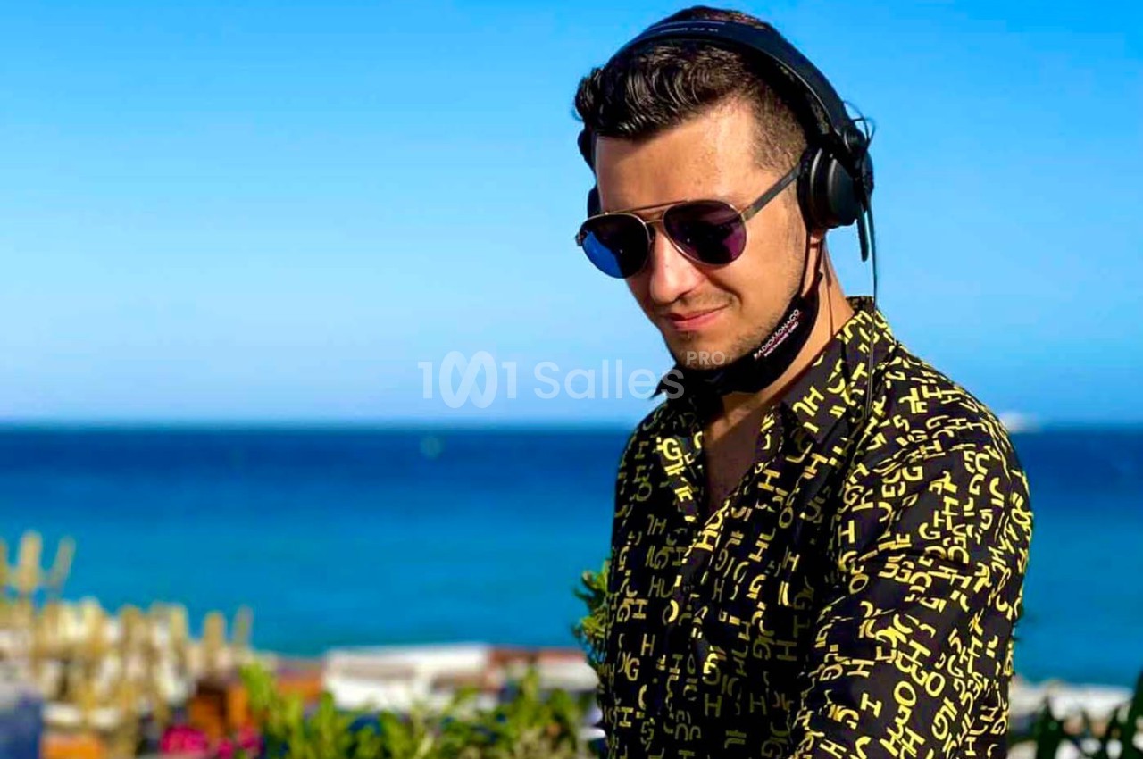 Un homme portant un casque audio et des lunettes de soleil mixe en extérieur près de la mer sous un ciel bleu.