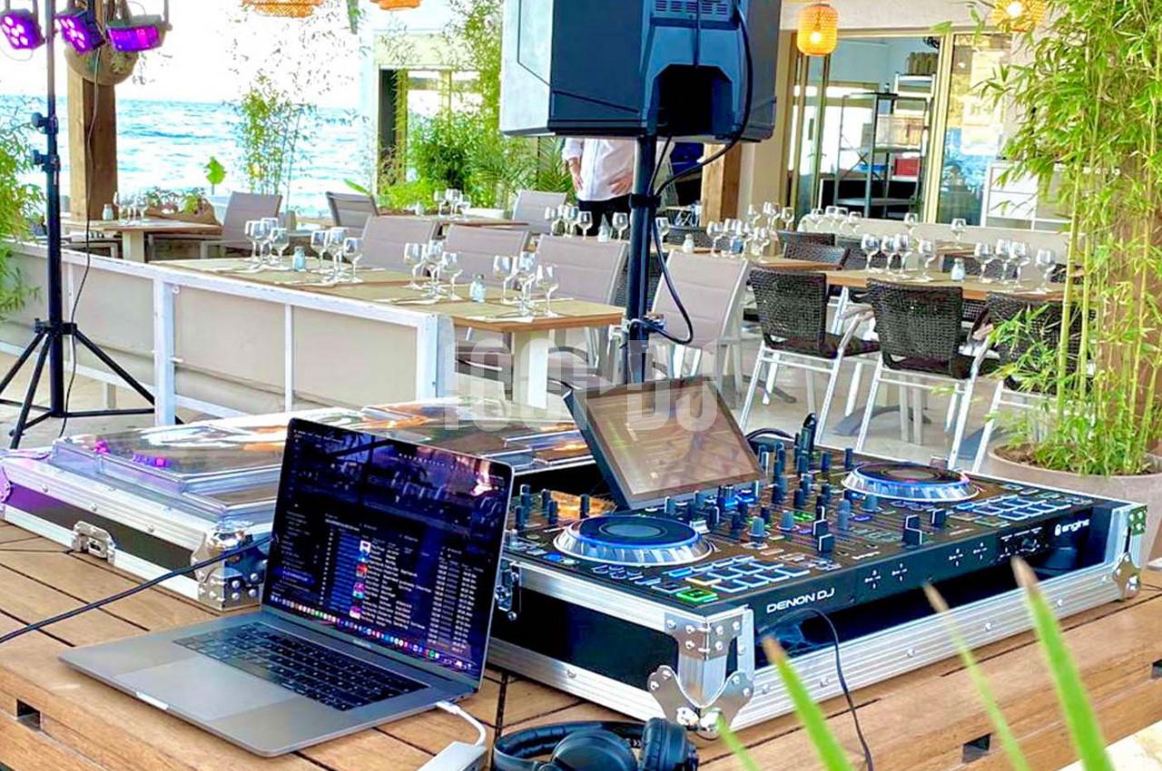 Table de mixage et ordinateur portable installés sur une terrasse en bord de mer, avec des tables dressées en arrière-plan.