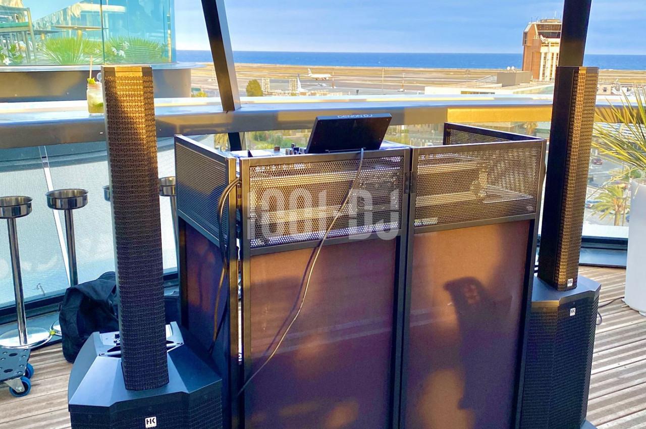 Poste de DJ avec équipement audio professionnel installé sur une terrasse offrant une vue sur la mer et des bâtiments.