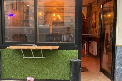 Miniature Location salle Paris 11 (Paris) - Bar de l'Hôtel de la Paix #14 Façade d'un café avec une table extérieure en bois, revêtement en gazon synthétique et intérieur éclairé visible.