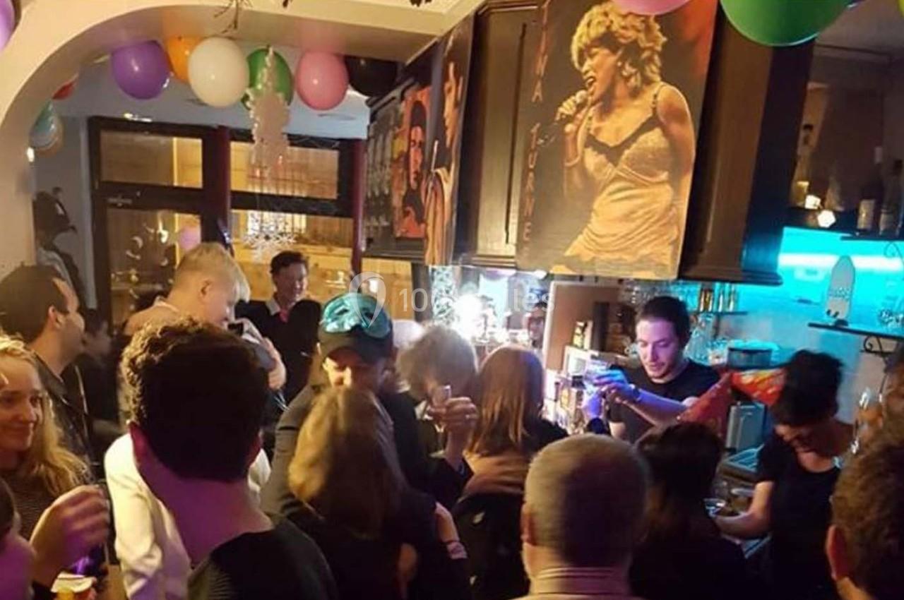 Groupe de personnes rassemblées dans un bar décoré de ballons, avec un barman servant des boissons.
