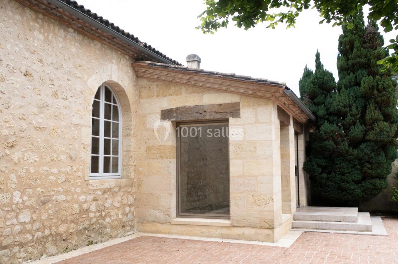Façade en pierre d'une maison avec une grande fenêtre, un arbre et une terrasse en briques au premier plan.