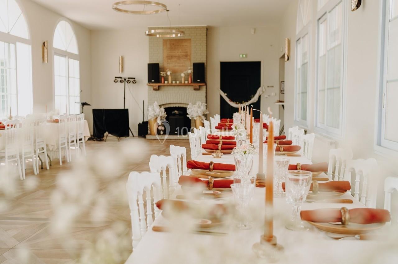Salle de réception lumineuse avec de longues tables décorées de bougies, serviettes rouges et vaisselle élégante.