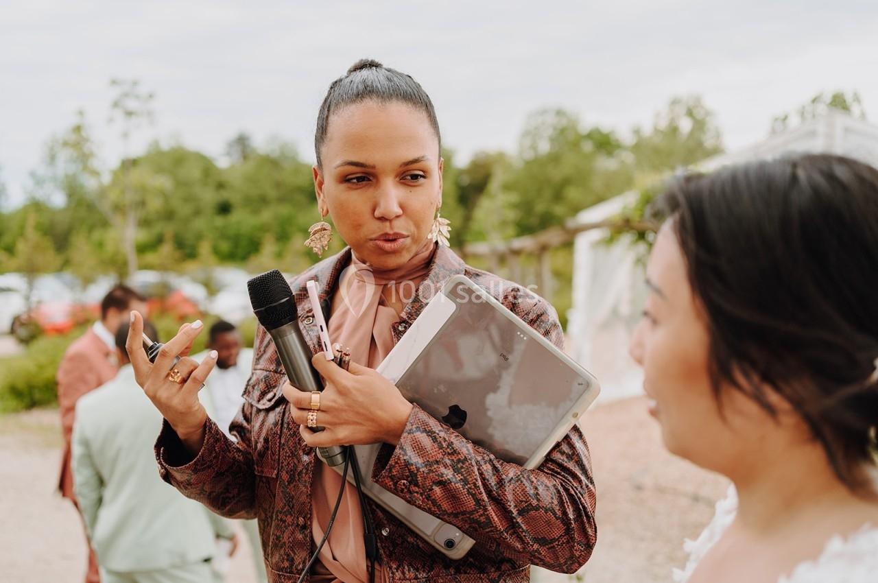 Une femme tenant un micro et une tablette parle à une autre personne en extérieur, avec des arbres en arrière-plan.