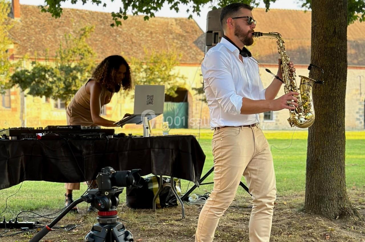 Un musicien joue du saxophone en plein air tandis qu'une DJ mixe sur une table équipée d'un ordinateur portable.