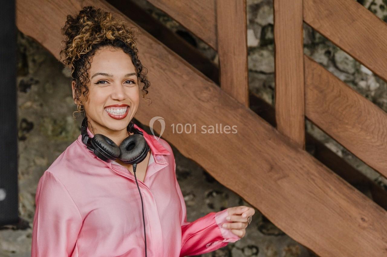 Femme souriante portant une chemise rose et un casque autour du cou, devant un escalier en bois.