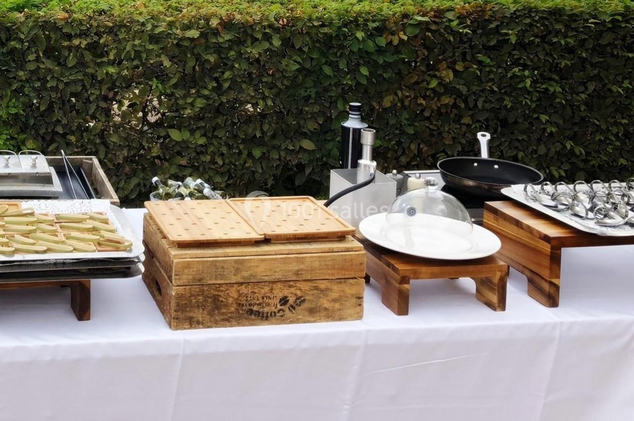 Table dressée en extérieur avec des plats, ustensiles de cuisine et présentoirs en bois devant une haie verte.