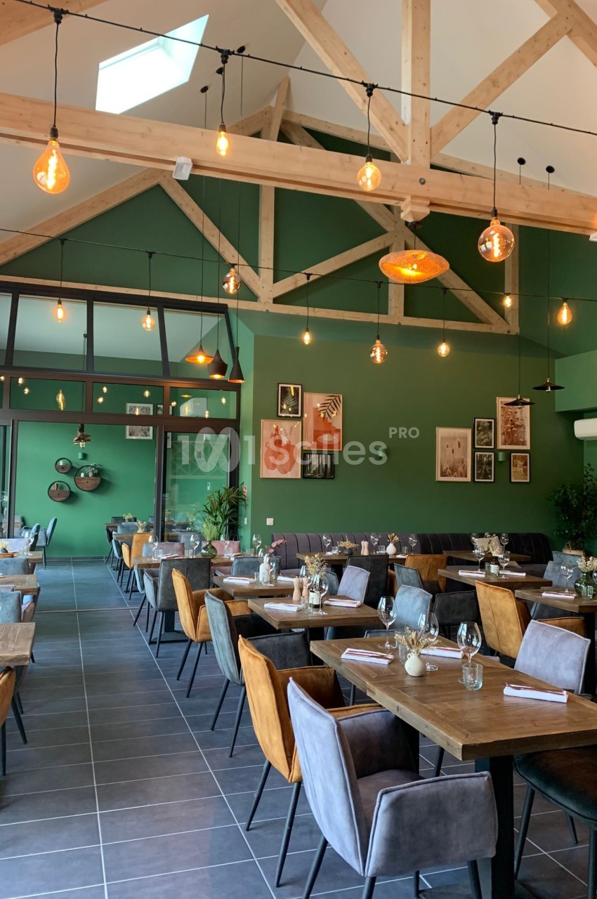 Salle de restaurant moderne avec murs verts, poutres apparentes, tables dressées et éclairage suspendu chaleureux.