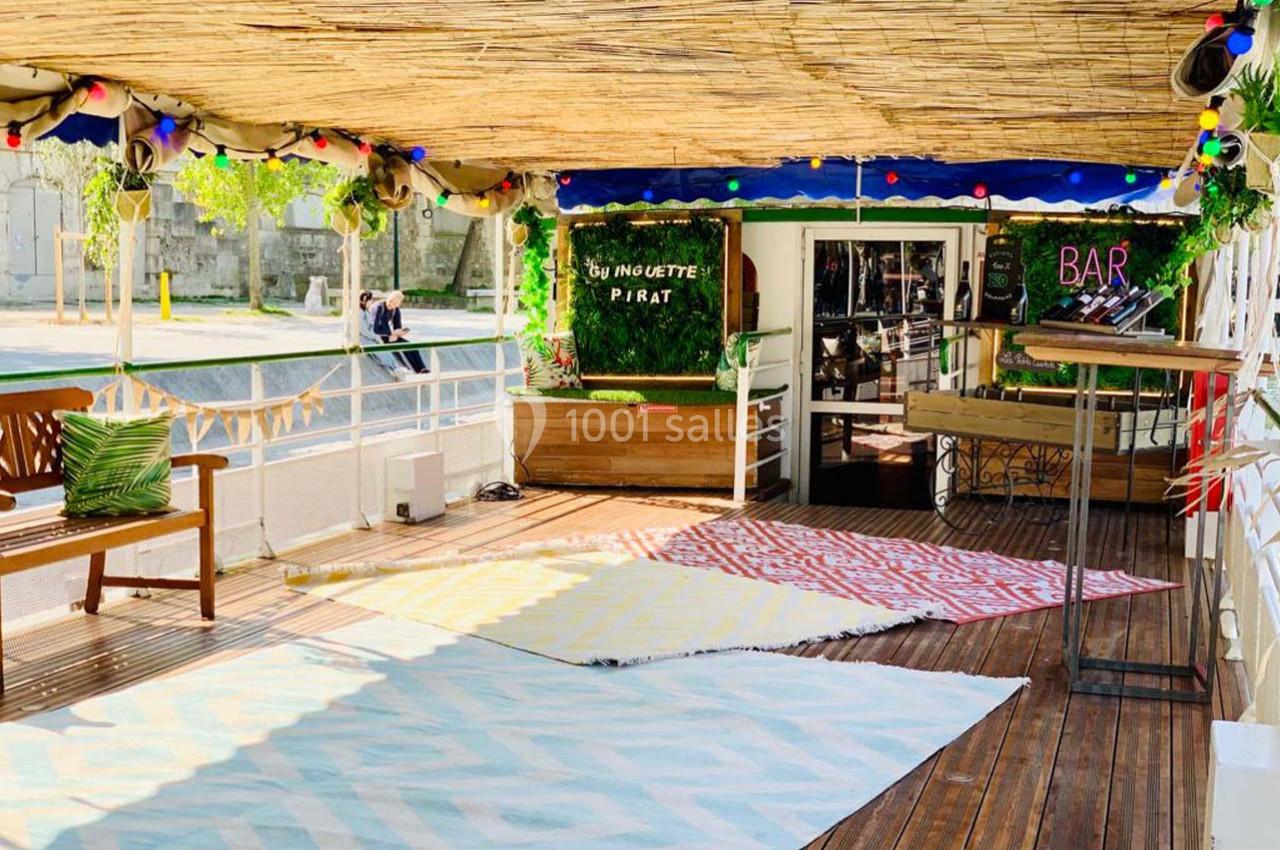 Terrasse en bois ombragée avec tapis colorés, banc en bois, décorations lumineuses et entrée d'un bar en arrière-plan.
