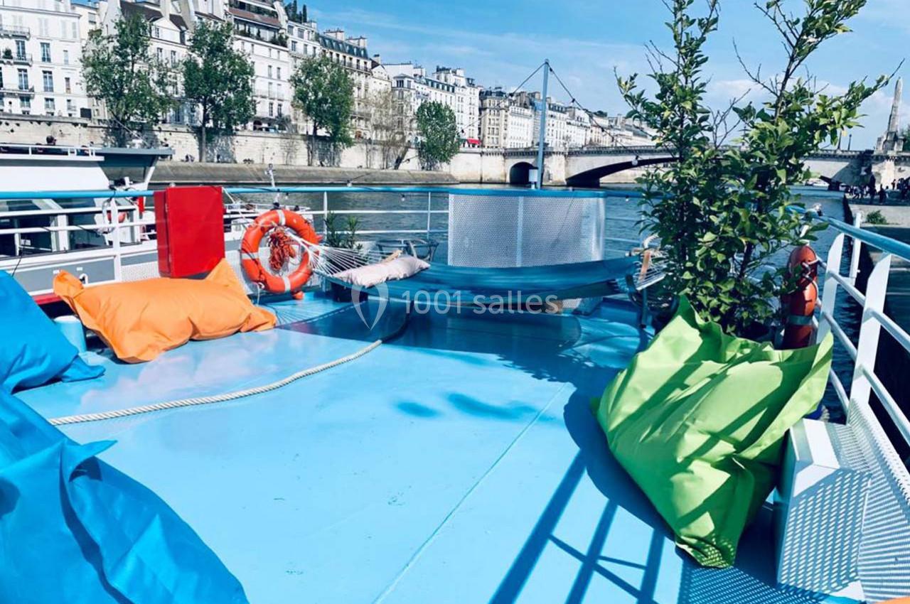 Terrasse d'un bateau avec poufs colorés, plantes en pot, bouée de sauvetage et vue sur des bâtiments en bord de Seine.