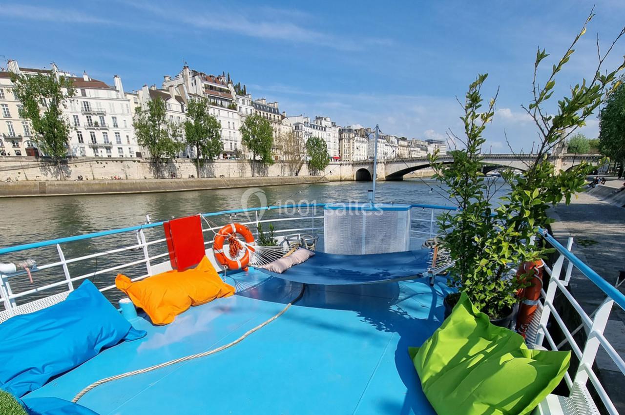 Pont d'un bateau avec coussins colorés, vue sur la Seine, des immeubles parisiens et un pont en arrière-plan.