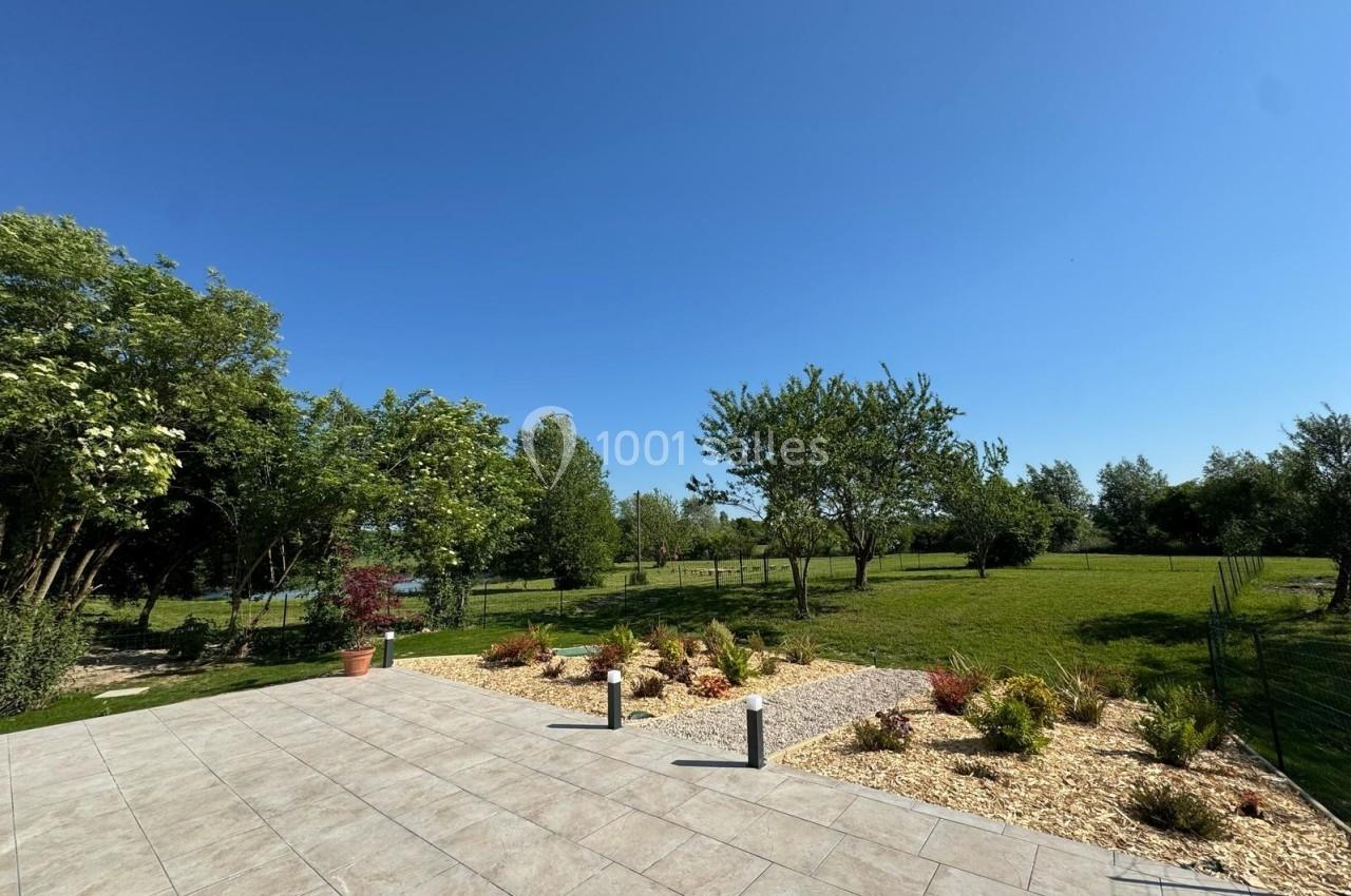 Vue d'une terrasse carrelée donnant sur un jardin arboré et une pelouse sous un ciel bleu dégagé.