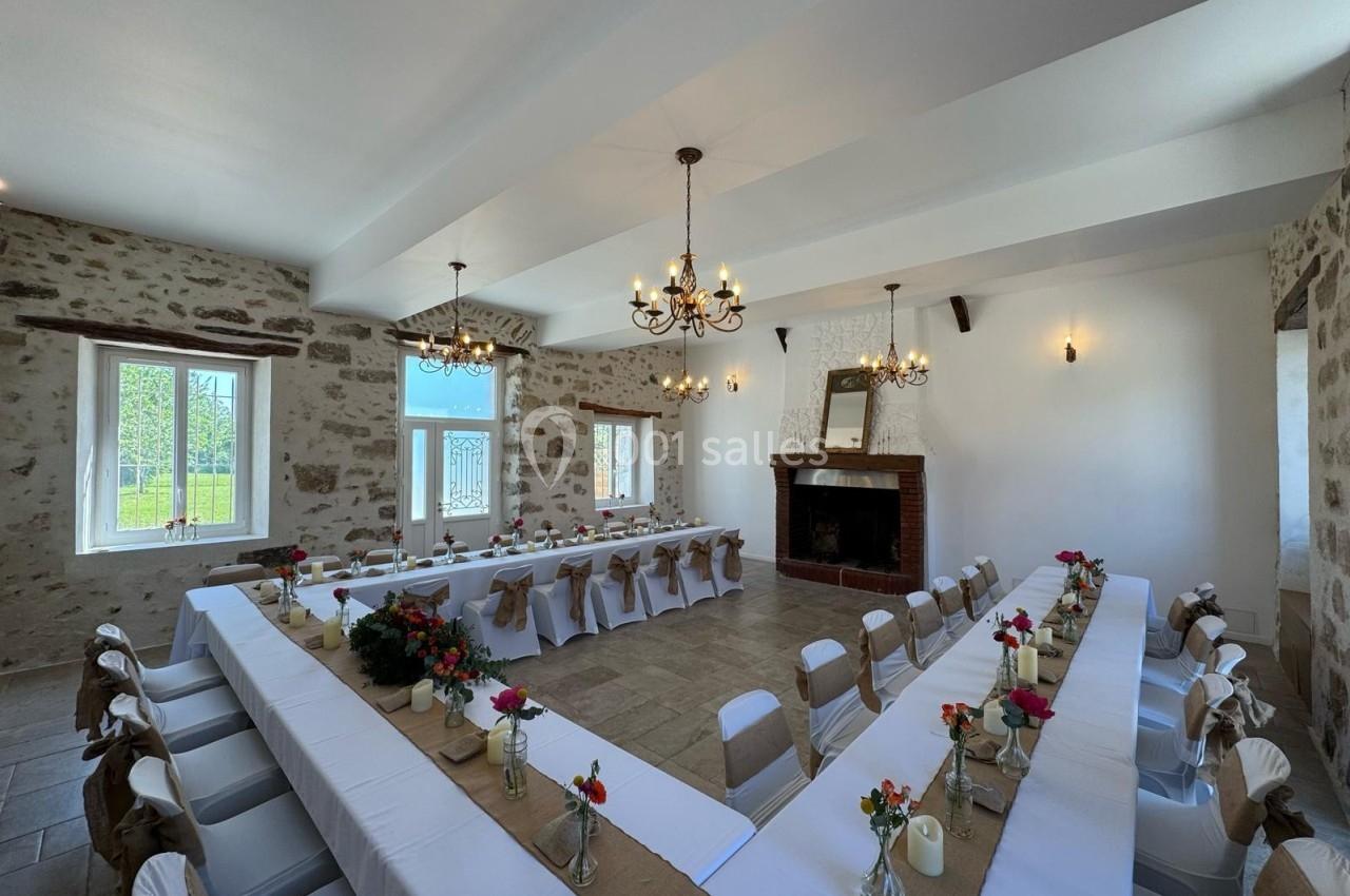 Salle de réception lumineuse avec tables en U décorées de nappes blanches, fleurs et bougies, murs en pierre apparente.