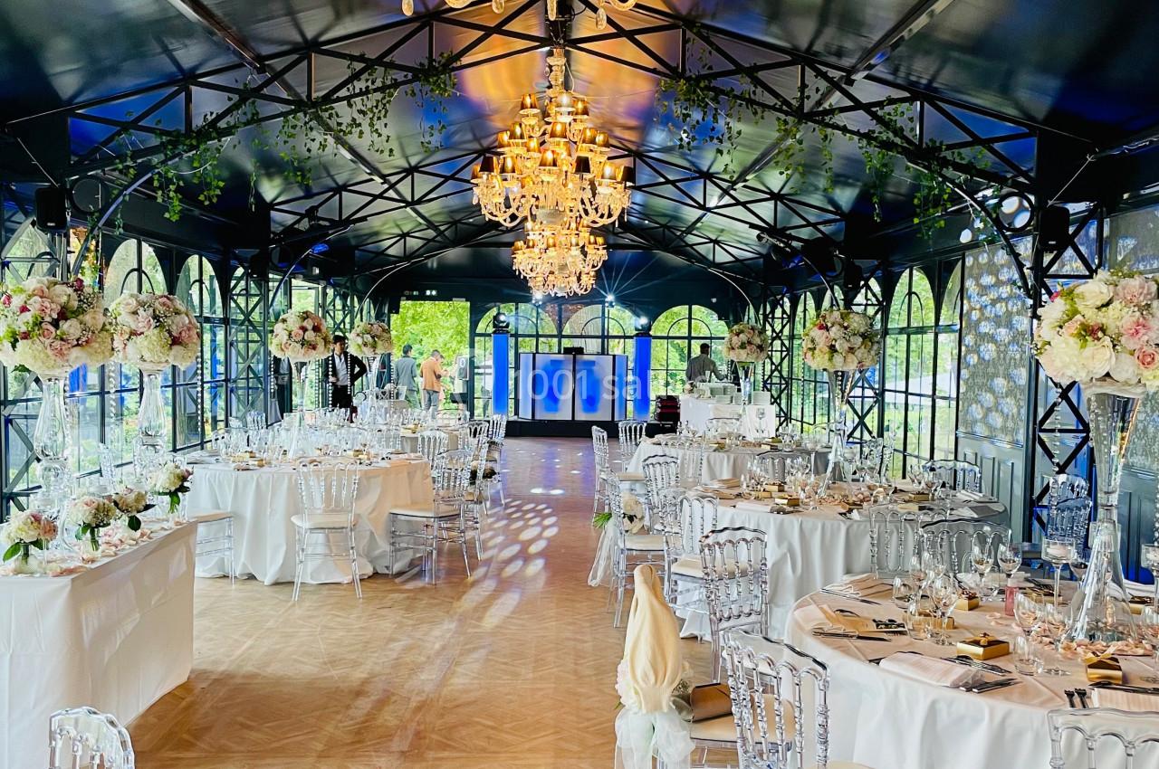 Salle de réception élégante avec tables décorées, chaises transparentes et lustres suspendus sous une verrière.