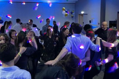 Des invités dansent joyeusement sous des lumières colorées dans une salle décorée pour une fête ou un mariage.