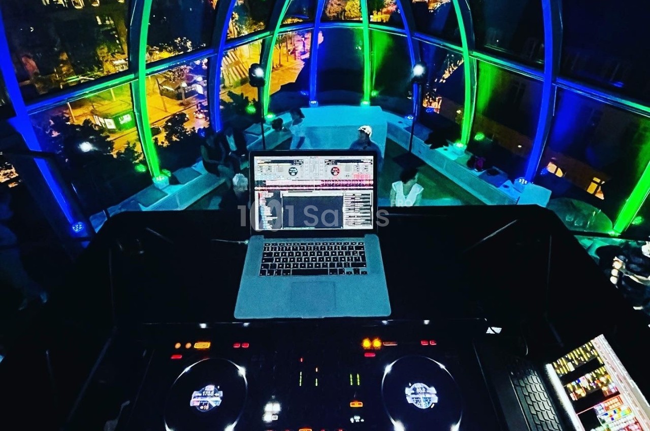 Poste de DJ avec platines et ordinateur, installé dans un espace vitré éclairé par des lumières colorées, vue nocturne en…